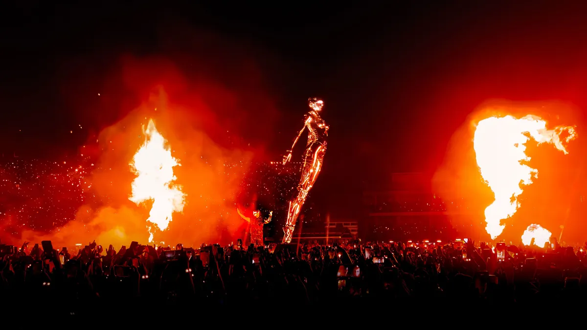 A stadium stage lit orange by two columns of flame, a small figure at the centre, a crowd of raised phones below