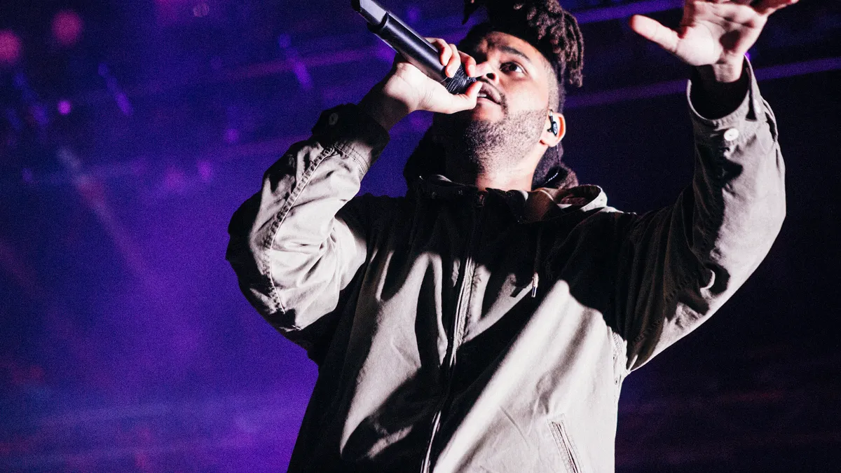 Abel Tesfaye, The Weeknd, singing into a handheld microphone with one arm raised, lit purple